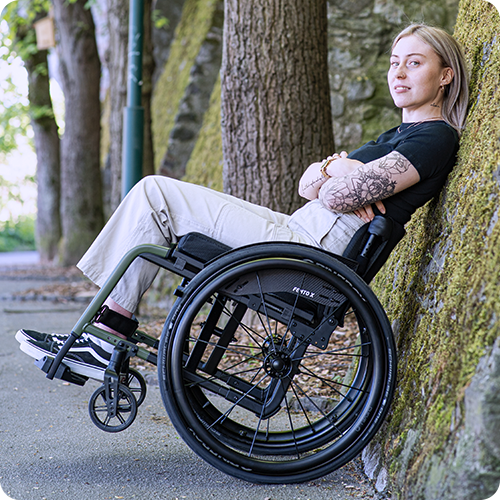 Eine junge Frau lehnt sich mit ihrem Rollstuhl an einen Steinmauer.