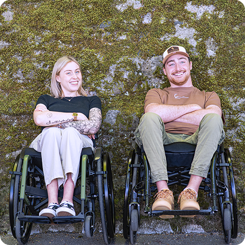 Eine junge Frau und ein junger Mann sitzen in Rollstühlen vor einer moosbewachsenen Steinmauer.
