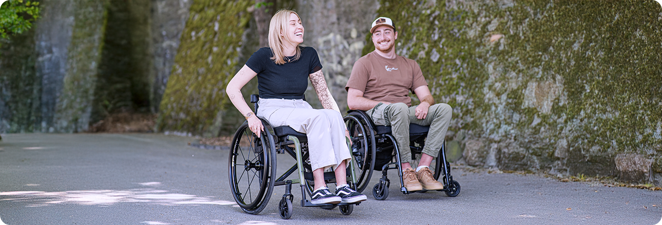 Eine junge Frau und ein junger Mann sitzen in Rollstühlen und gucken sich an.