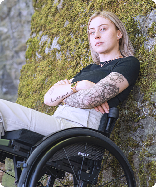Eine junge Frau sitzt in ihrem Rollstuhl an einer Steinmauer.